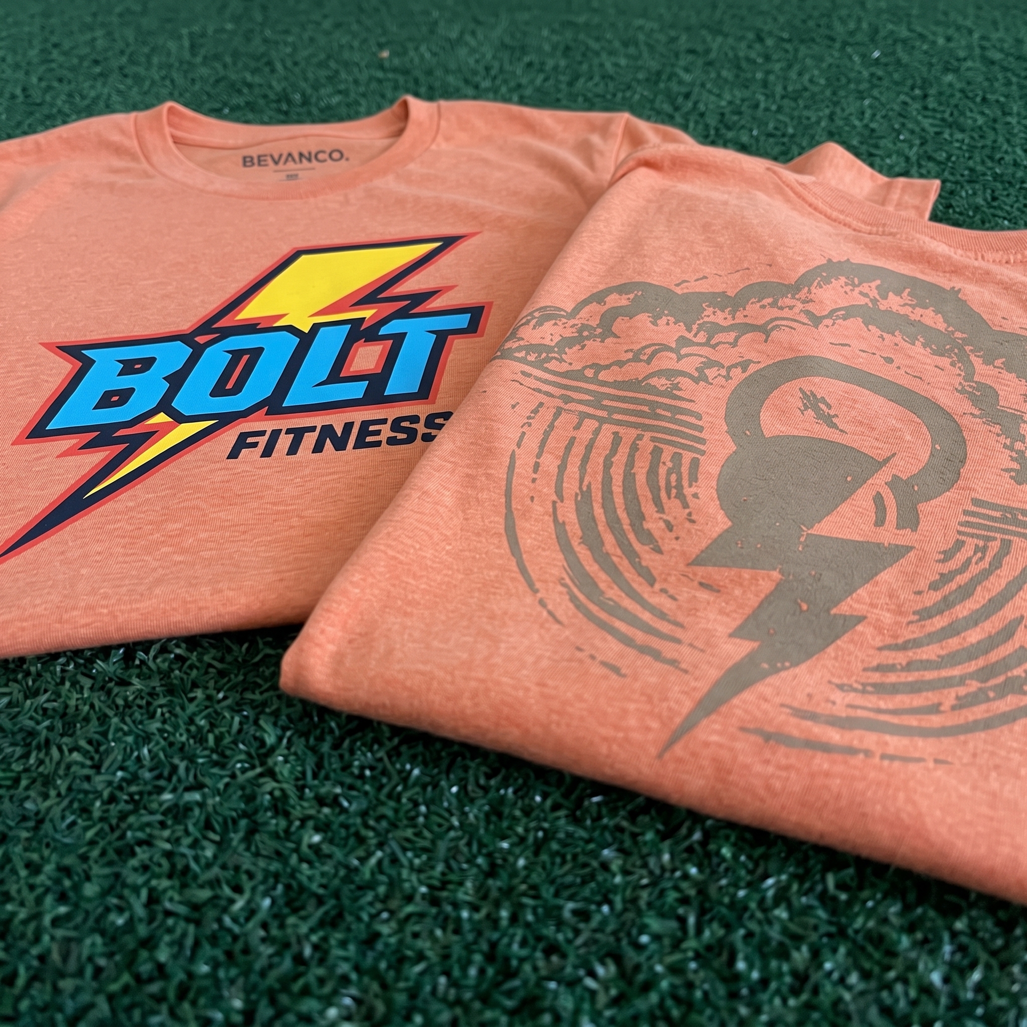 Two orange Bolt Fitness tees on green turf with full-color front logo and tonal kettlebell lightning back graphic