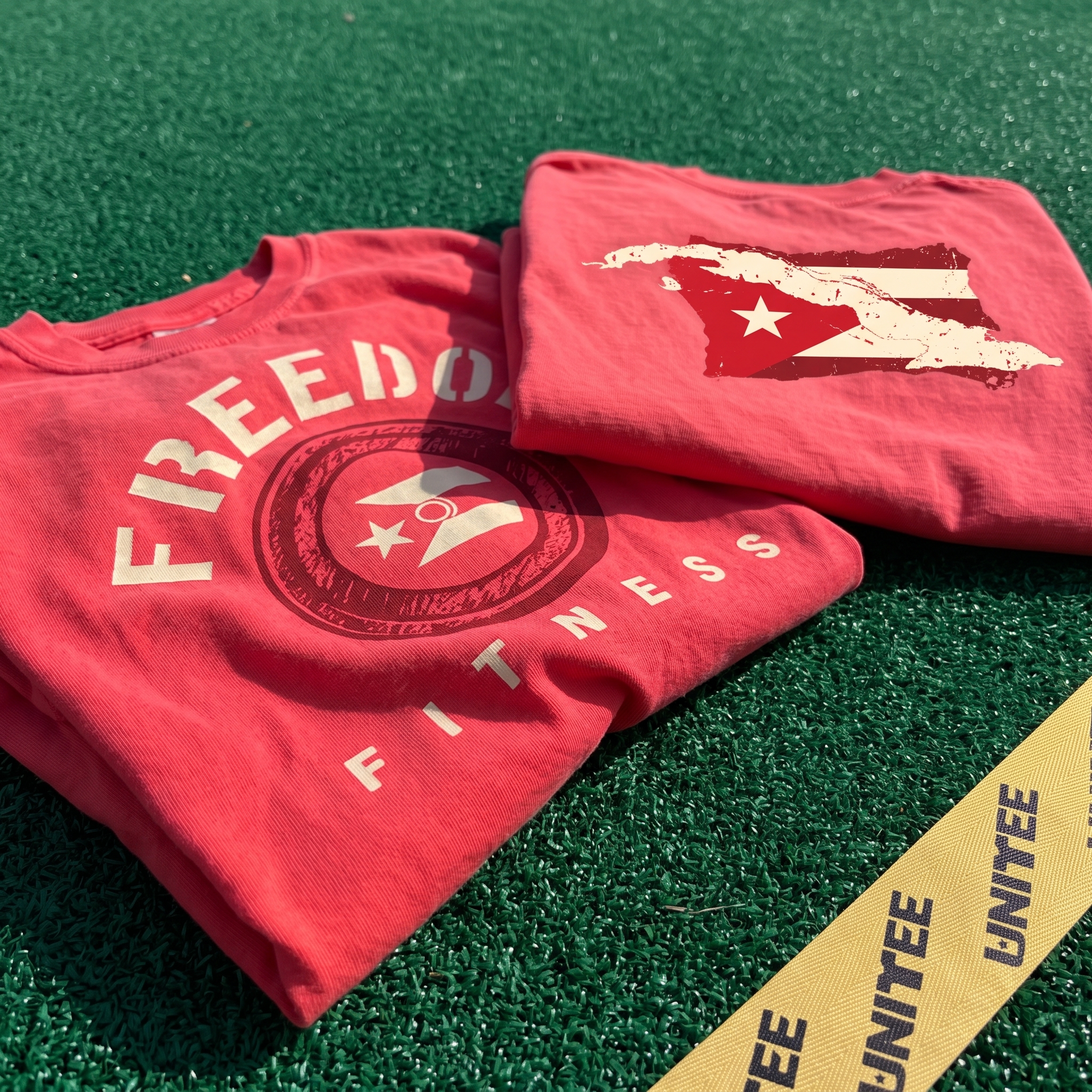 Two coral Freedom Fitness tees on green turf with circular stamp graphic and Cuban flag shaped back print