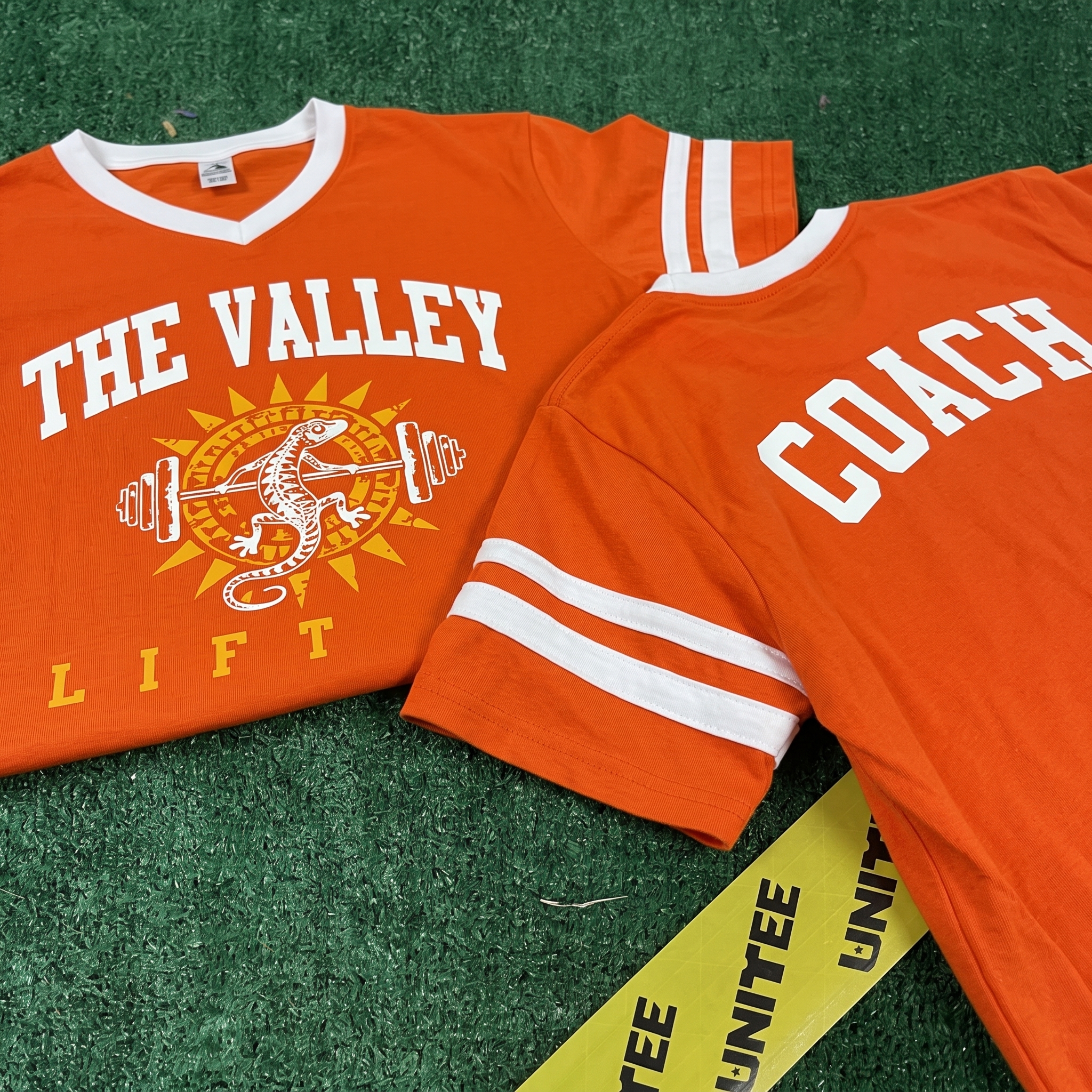 Orange The Valley Lift Co jersey-style tee with coach back lettering, sleeve stripes, lizard sunburst logo, and white type