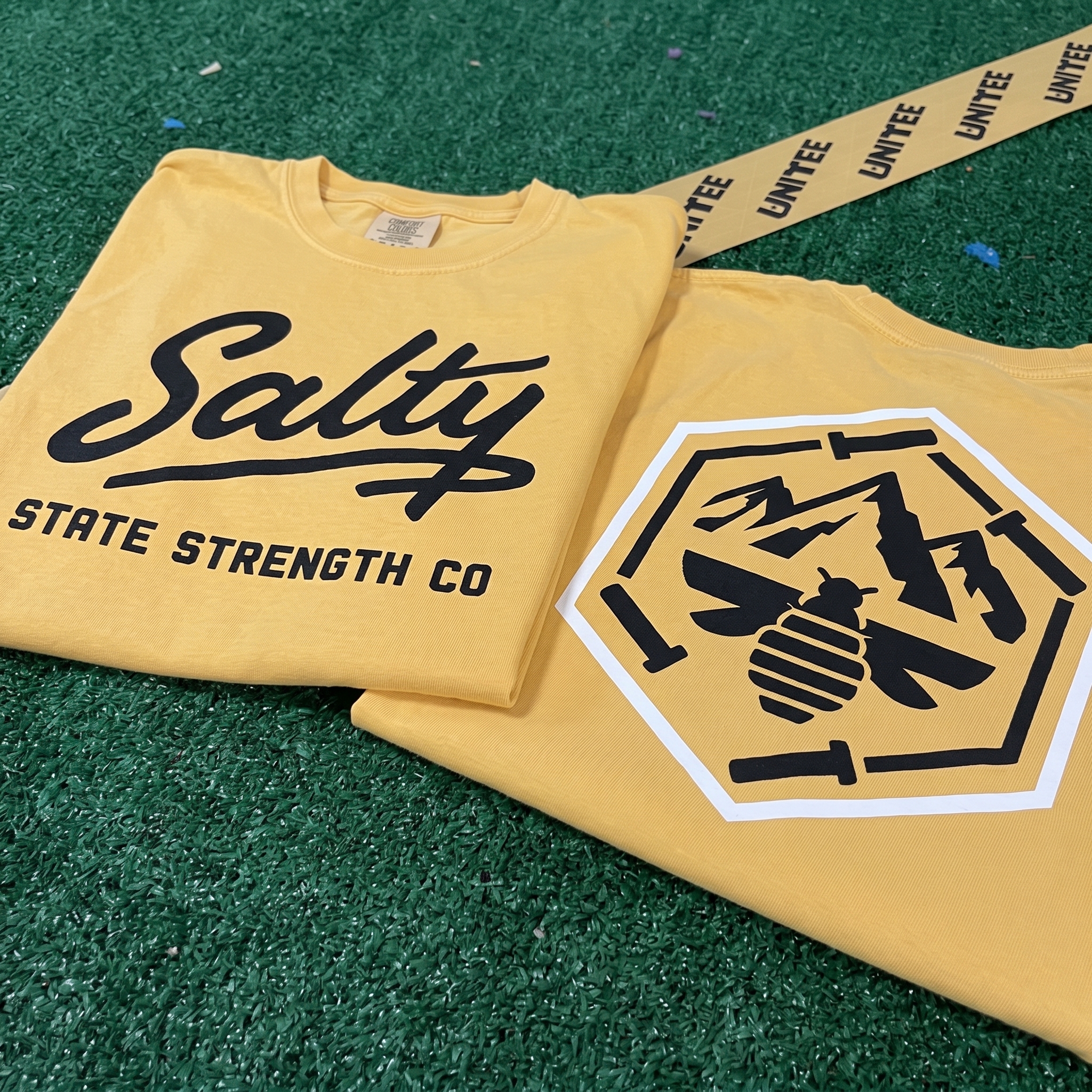 Yellow Salty State Strength Co. tees on turf with front script logo and back mountain bee badge