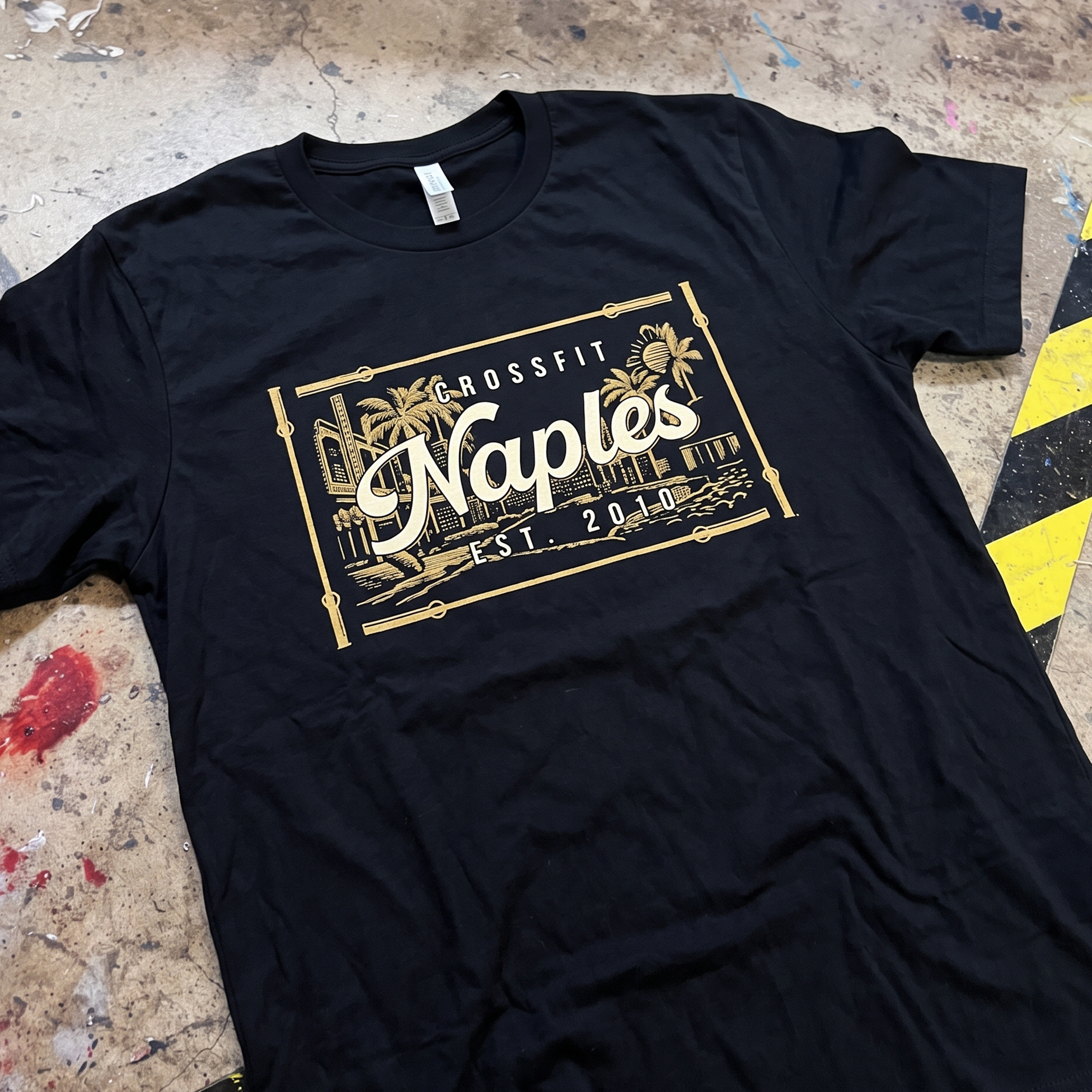 Black CrossFit Naples tee with a gold and cream framed Naples skyline graphic.