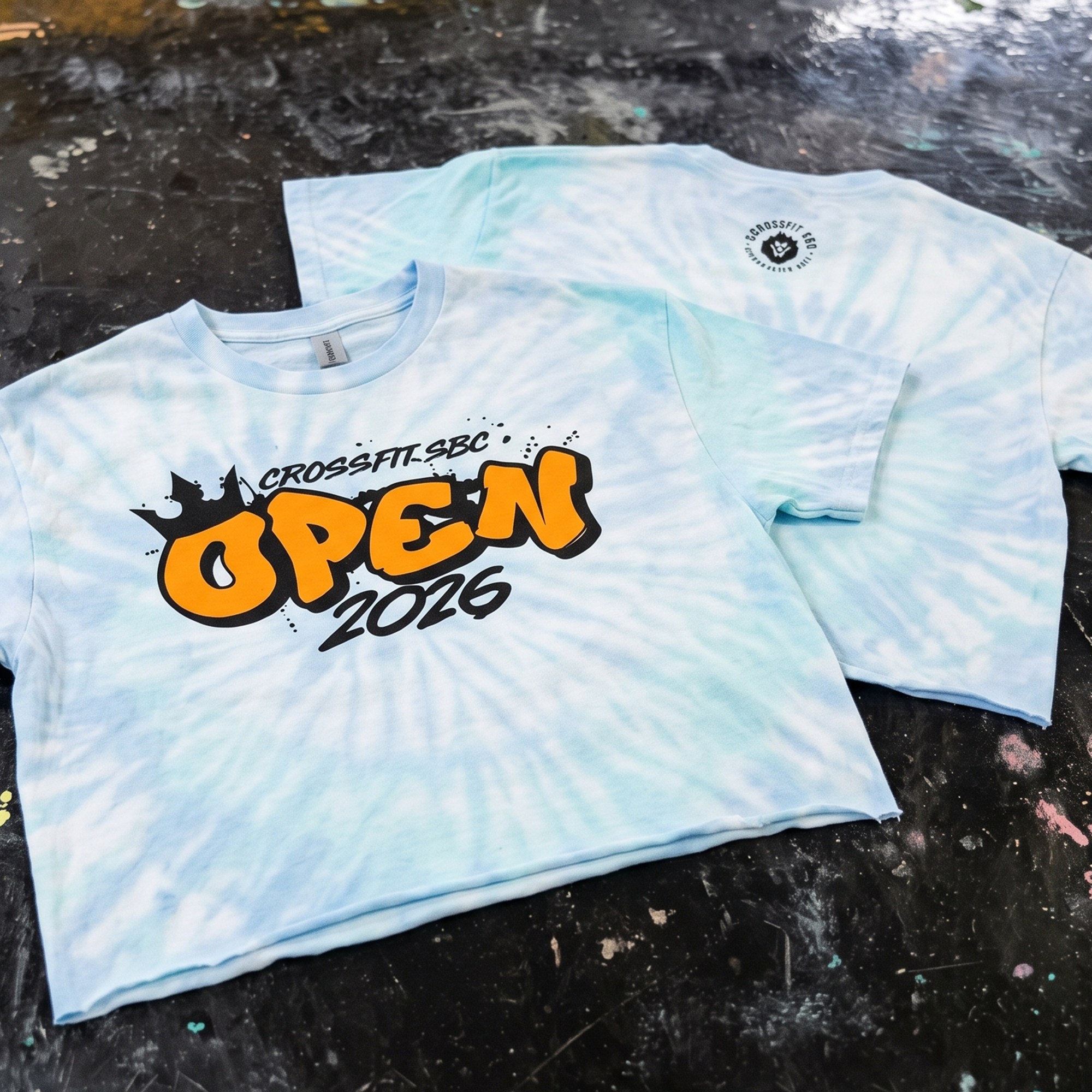 Light blue tie-dye tee with black and orange CrossFit SBC Open 2026 artwork and a small upper-back logo.