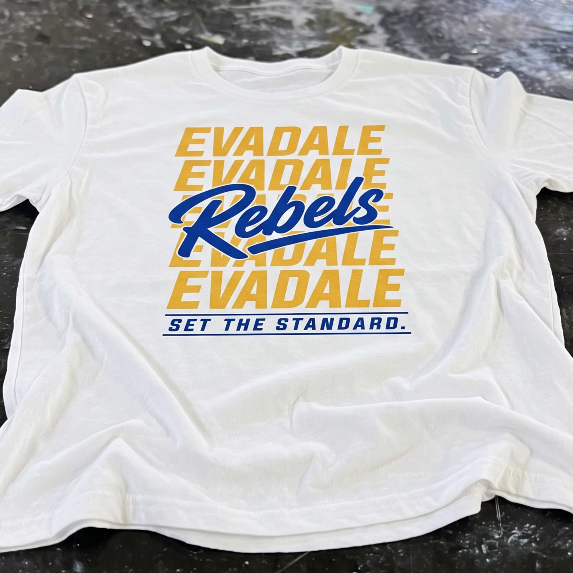White T-shirt with repeated gold Evadale lettering, blue Rebels script, and Set the Standard text.