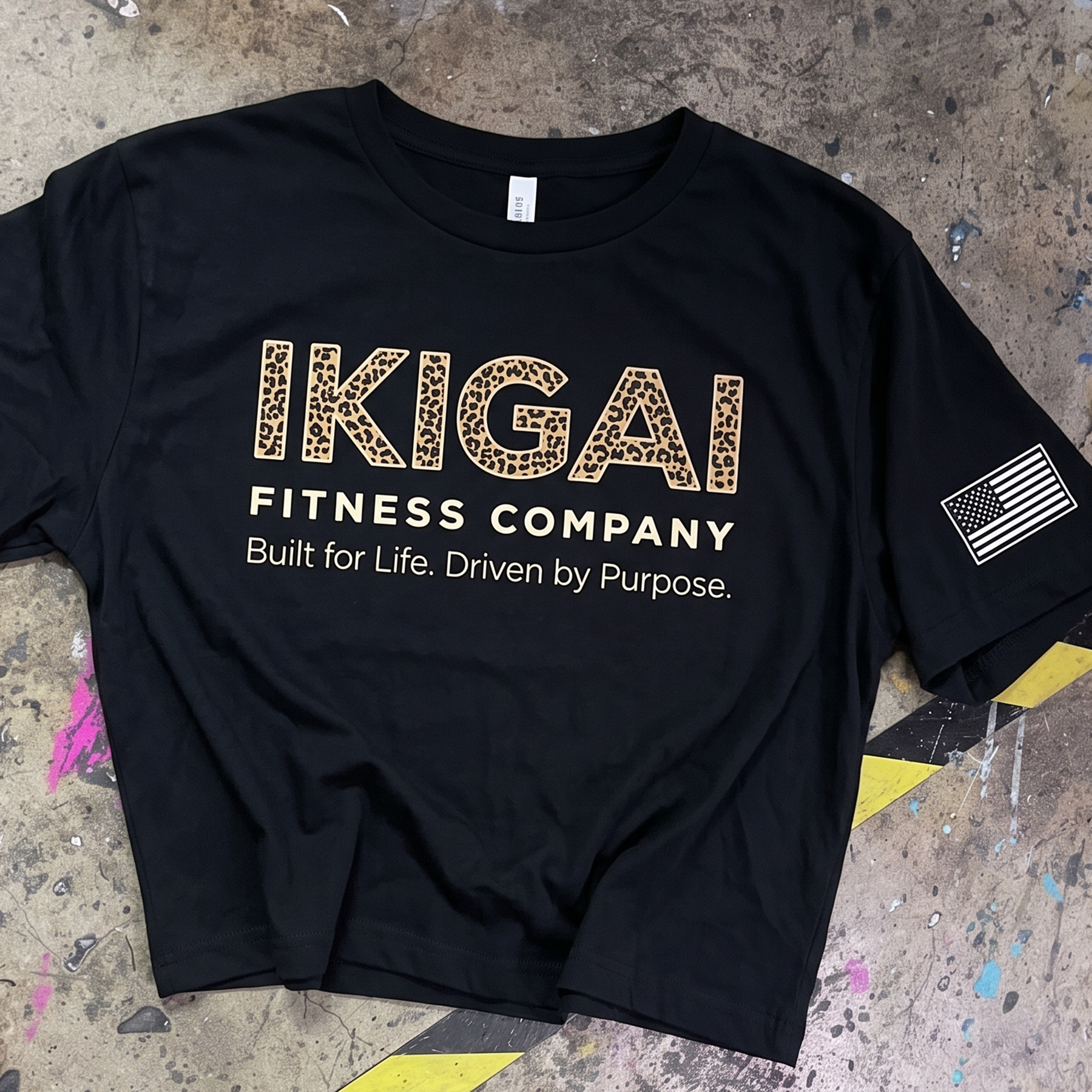 Black Ikigai Fitness Company T-shirt with leopard-print logo, cream text, and an American flag sleeve print.