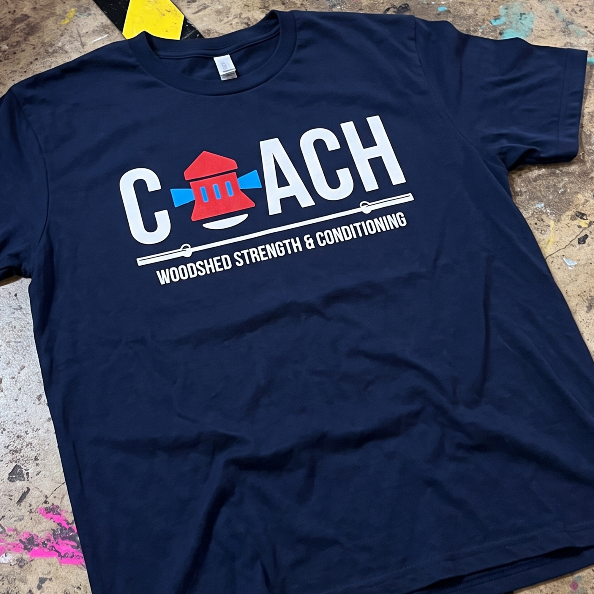 Navy T-shirt with white coach lettering, red-and-blue icon, and barbell graphic for Woodshed Strength & Conditioning.