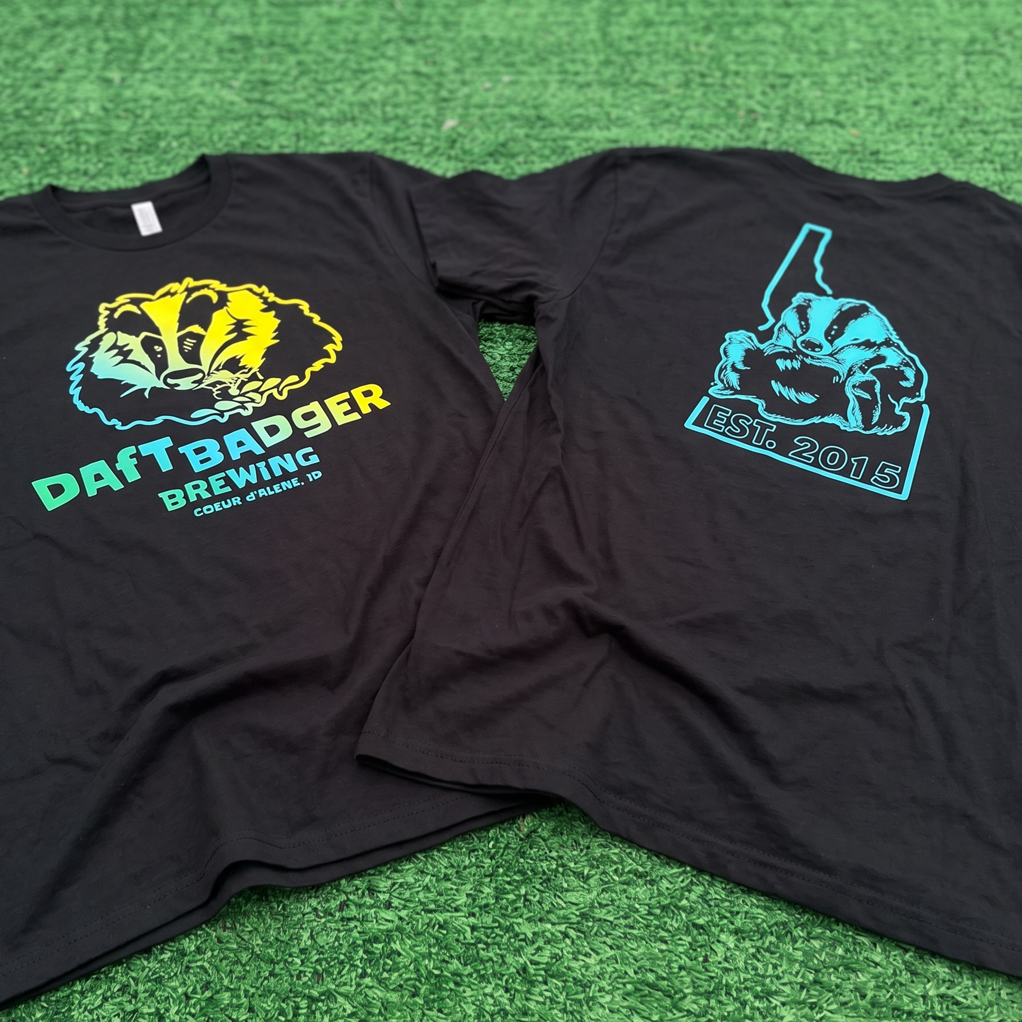 Two black Daft Badger Brewing T-shirts with colorful badger graphics on green turf.